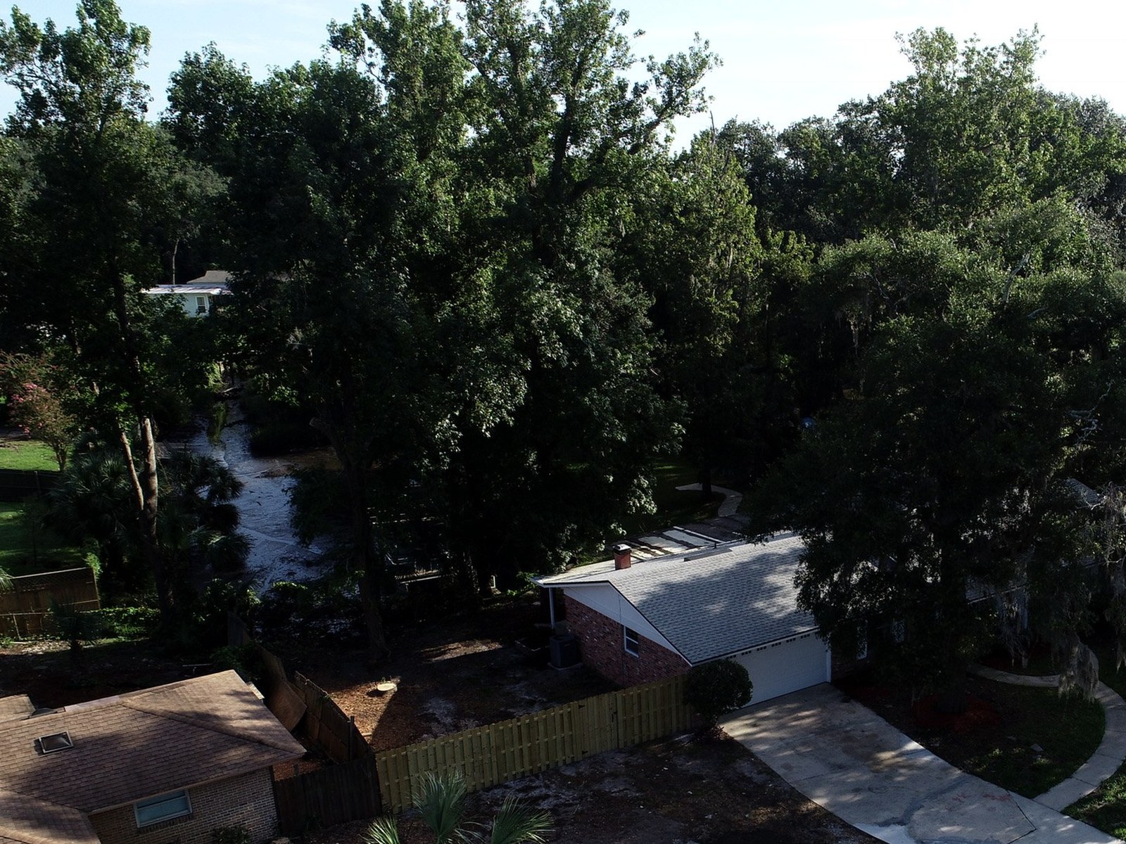Aerial view of a single story brick home with a gray shingle roof and white garage door beneath tall shade trees, with a wooden fence, a neighboring roof and a small creek through the trees, Jacksonville, Duval County, Florida